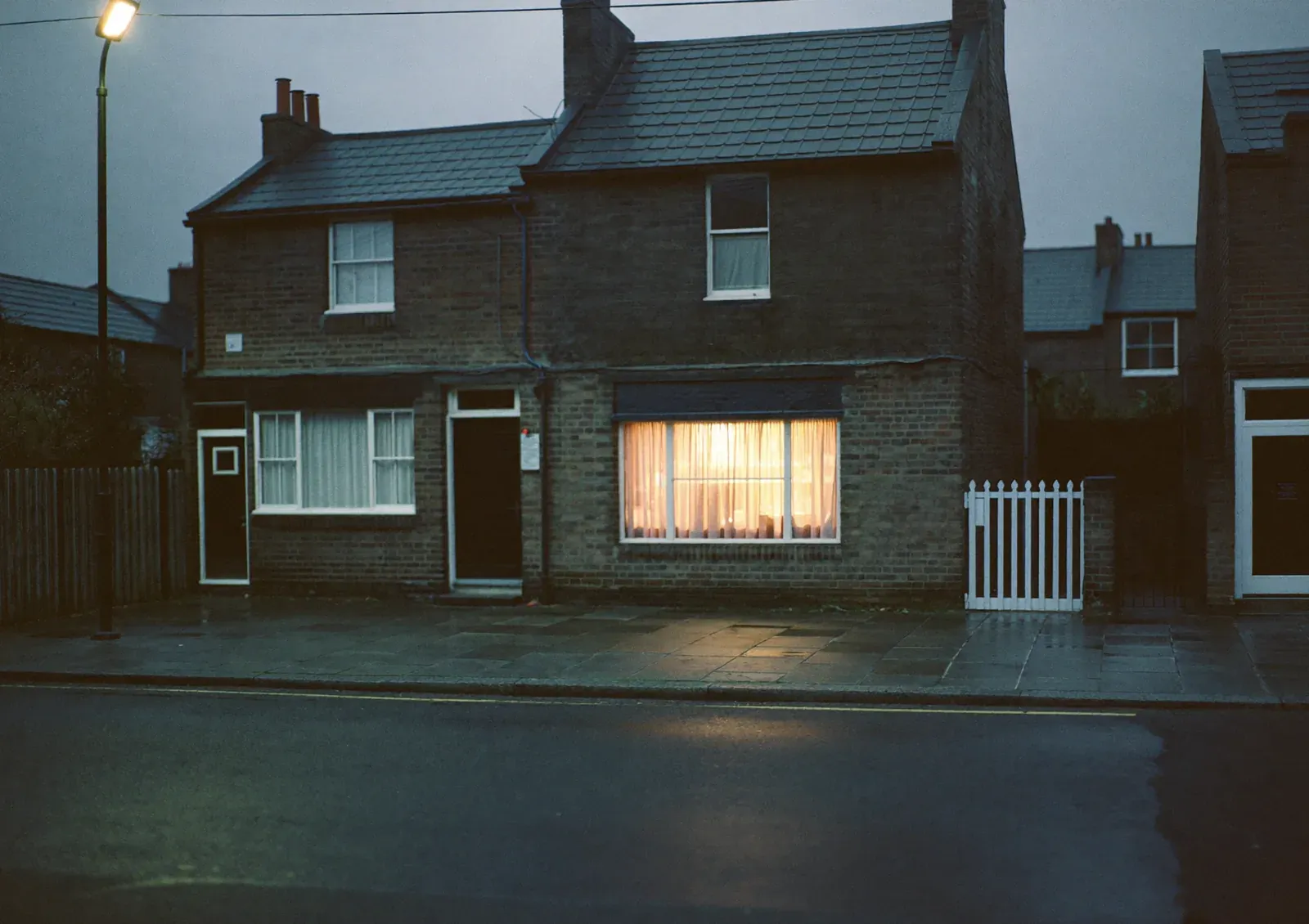 A British residential street at dusk with one front room lit behind the curtains.